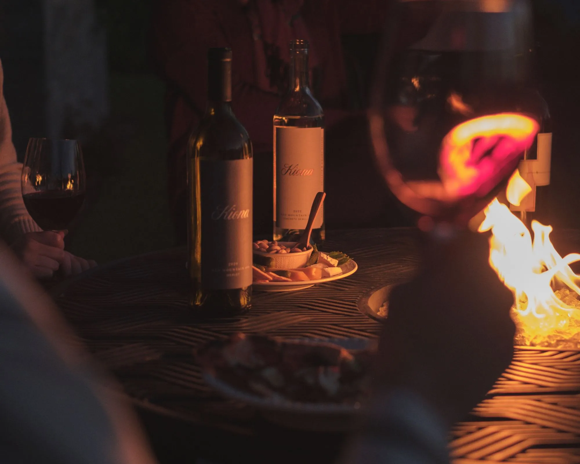 Two bottles on a table next to a fire with an arm holding a wine glass in the foreground